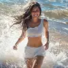 HDR photo of woman walks in the surf at a beach, long brunette hair blowing in the wind, laughing at the viewer, water splashing, bright sunlight, wearing a white t-shirt, white shorts . High dynamic 