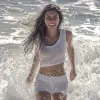 HDR照片中，一名女子在海滩的海浪中行走，深色长发在风中飘扬，嘲笑观众，水花四溅，明亮的阳光，穿着白色T恤、白色短裤。动态范围高、生动、细节丰富、阴影和亮点清晰、逼真、强烈、对比度增强、高度细致