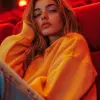 photography, vast emptiness of a movie theater serves as the backdrop for a moment of carefree repose, woman lounges across the seats, a picture of relaxed ease in an oversized orange color hoody and 