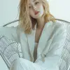 创造一位年轻的韩国女模特的形象，拥有齐肩金发，在极简主义环境中拍摄了一张引人注目的当代照片，她躺在一把建筑风格的哑光银色金属椅子上，设计有大胆的线性框架，模特优雅地穿着白色西装外套和裤子，她的姿势放松但充满意图，舒适地向后靠，双腿随意地搭在椅子的侧面，她的手臂沿着顶部松垮垮地靠在上面，头稍微向后倾斜，给人一种轻松和自信的感觉，背景是朴素、明亮的白色，凸显了椅子'的刚性结构和她柔软、流畅的姿