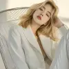 创造一位年轻的韩国女模特的形象，拥有齐肩金发，在极简主义环境中拍摄了一张引人注目的当代照片，她躺在一把建筑风格的哑光银色金属椅子上，设计有大胆的线性框架，模特优雅地穿着白色西装外套和裤子，她的姿势放松但充满意图，舒适地向后靠，双腿随意地搭在椅子的侧面，她的手臂沿着顶部松垮垮地靠在上面，头稍微向后倾斜，给人一种轻松和自信的感觉，背景是朴素、明亮的白色，凸显了椅子'的刚性结构和她柔软、流畅的姿